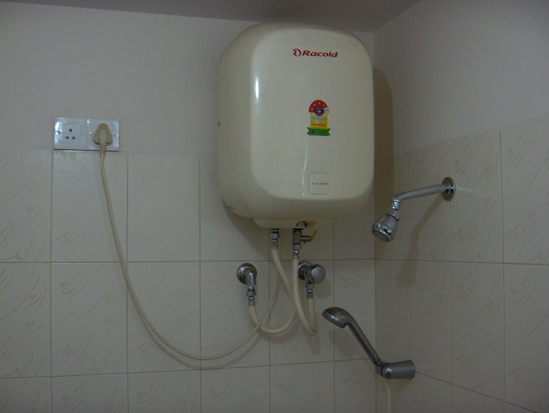En vit elektrisk varmvattenberedare av märket Racold monterad på en badrumsvägg med anslutna rör och duschdetaljer.